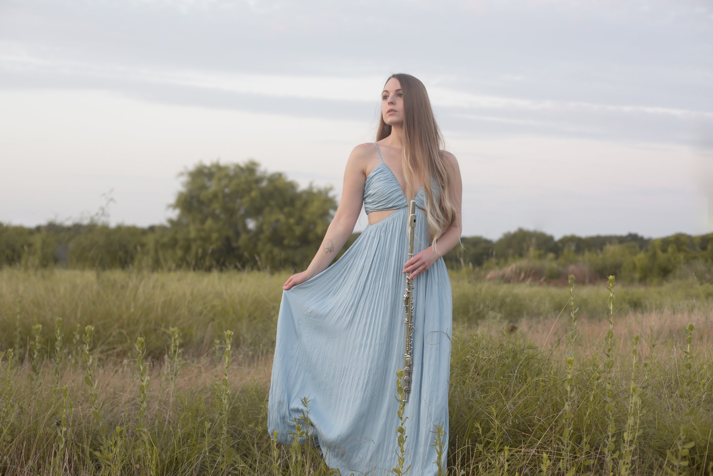 Leanna Ginsburg standing in a field at dusk, holding her flute, wearing a pale blue dress.